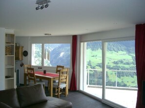 Ein Zimmer mit Esstisch, Stühlen, einer Couch und großen Fenstern mit Blick auf eine Berglandschaft.