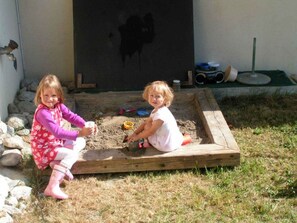 Dos niños jugando en un arenero con un marco de madera, rodeado de rocas y césped.
