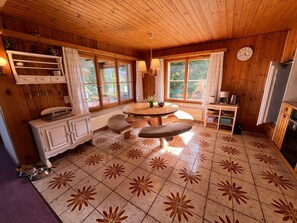 Interior de cabaña con techo de madera, una mesa de comedor redonda, una zona de cocina con refrigerador y un ventanal grande.