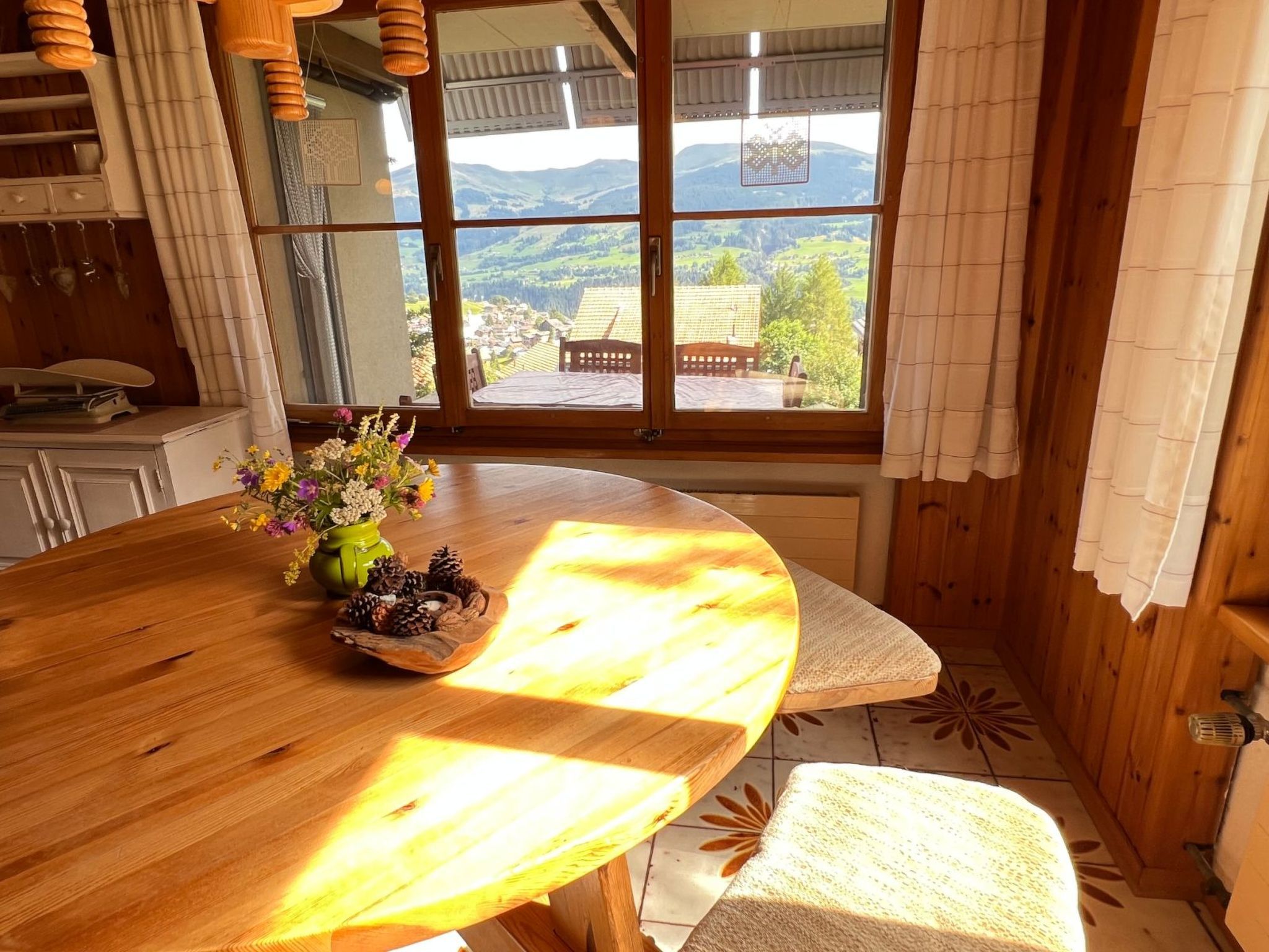 Ein Holztisch mit einer Blumenvase und Kiefernzapfen, eine Bank und Blick auf ein Dorf durch große Fenster.