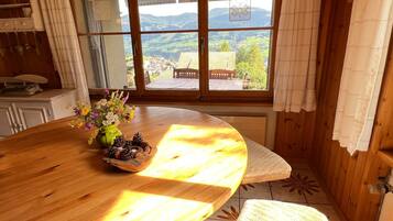 Ein Holztisch mit einer Blumenvase und Kiefernzapfen, eine Bank und Blick auf ein Dorf durch große Fenster.
