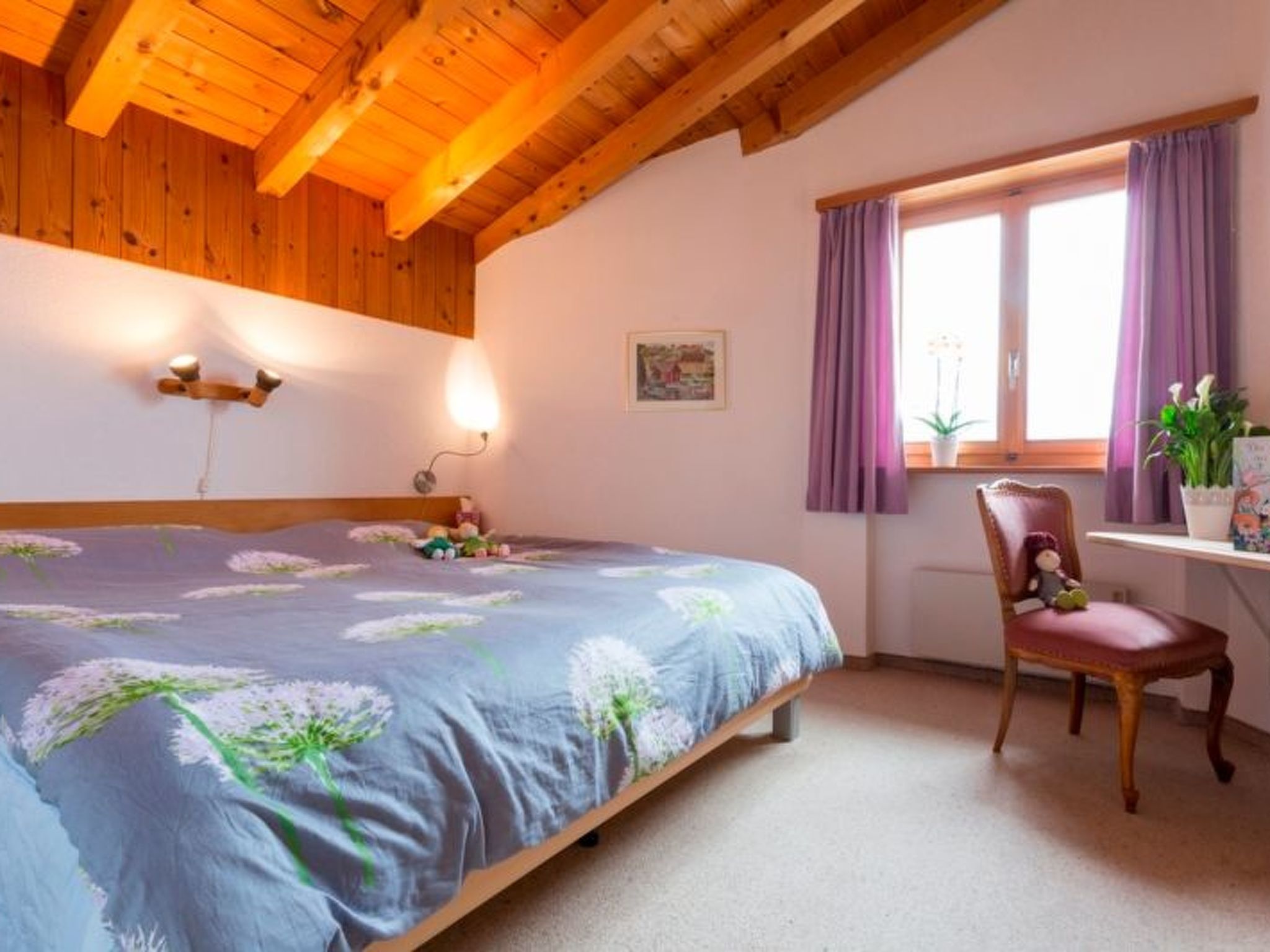 Un dormitorio con cama, escritorio, silla, ventana con cortinas y lámpara de pared.
