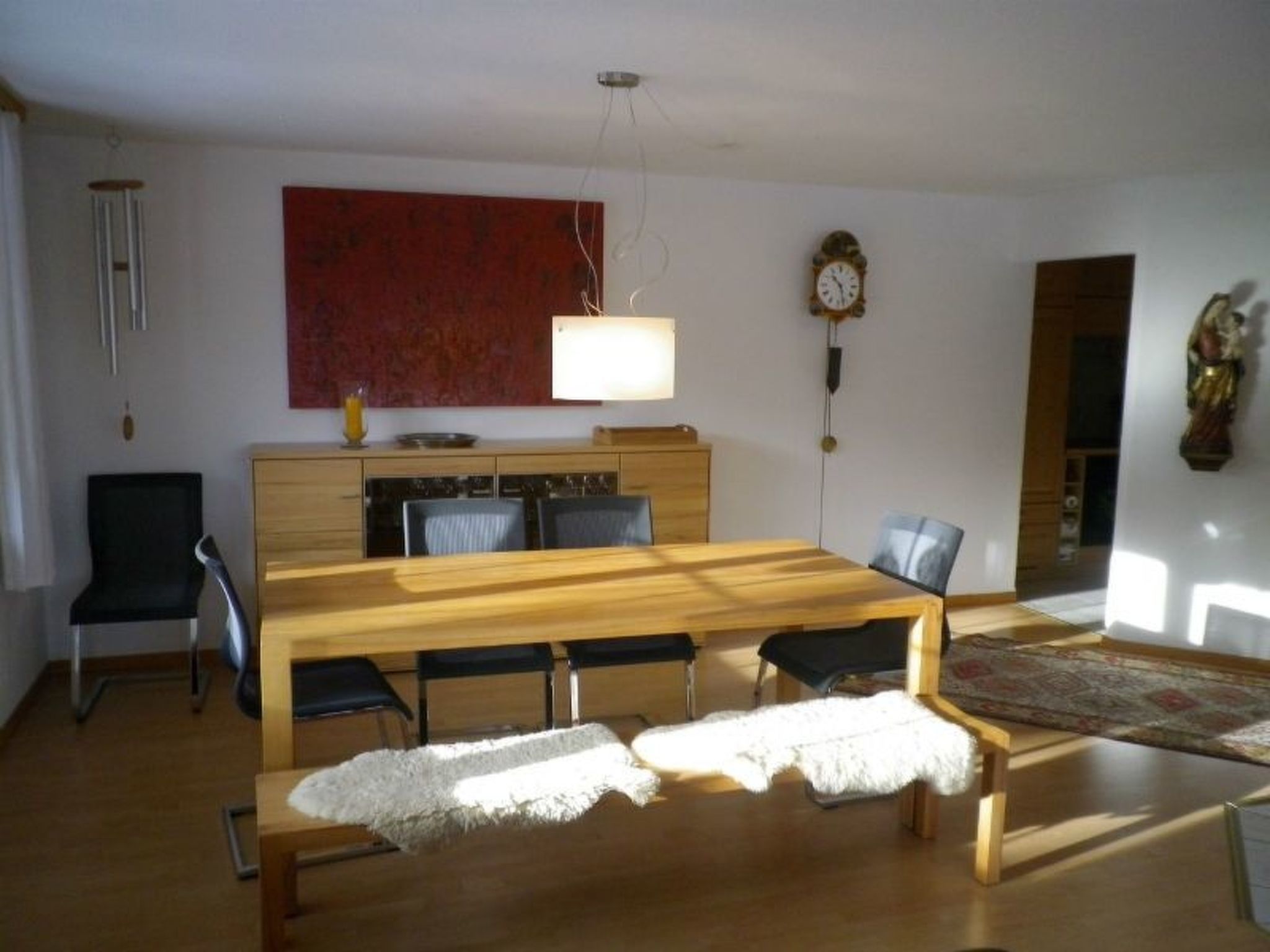 A dining area with a wooden table and benches, a kitchen in the background, and a large red painting on the wall.