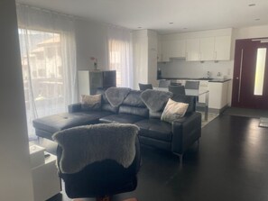 Uma sala de estar moderna com um sofá de couro escuro, uma poltrona combinando e uma área de jantar com cozinha ao fundo.
