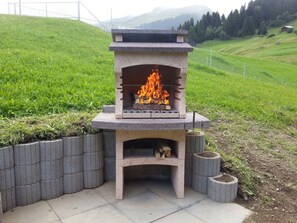 A stone-built outdoor grill with a fire burning inside, surrounded by a concrete base and a stone wall.