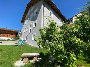 Ein Gebäude mit einer grauen Fassade und zahlreichen Fenstern, ein kleiner Gartenbereich mit einer Bank und Stühlen sowie ein Baum im Vordergrund.
