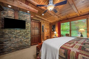 A bedroom with a bed, a ceiling fan, a TV, a stone wall, wooden paneling, and a window with blinds. - Swan Way Mountain Retreat (Lake Lure)