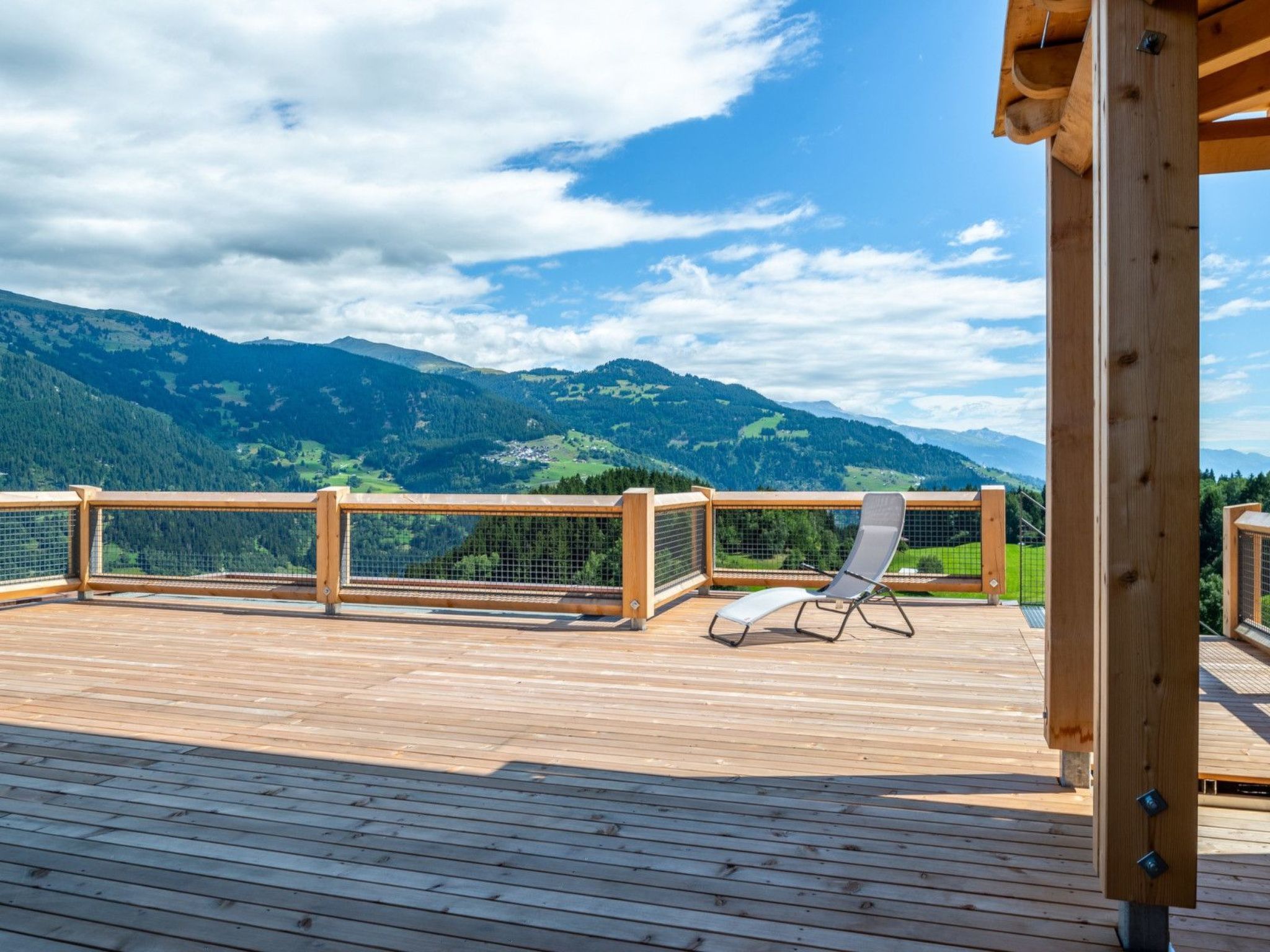 A wooden deck with a lounge chair, a view of green mountains, and a clear sky.