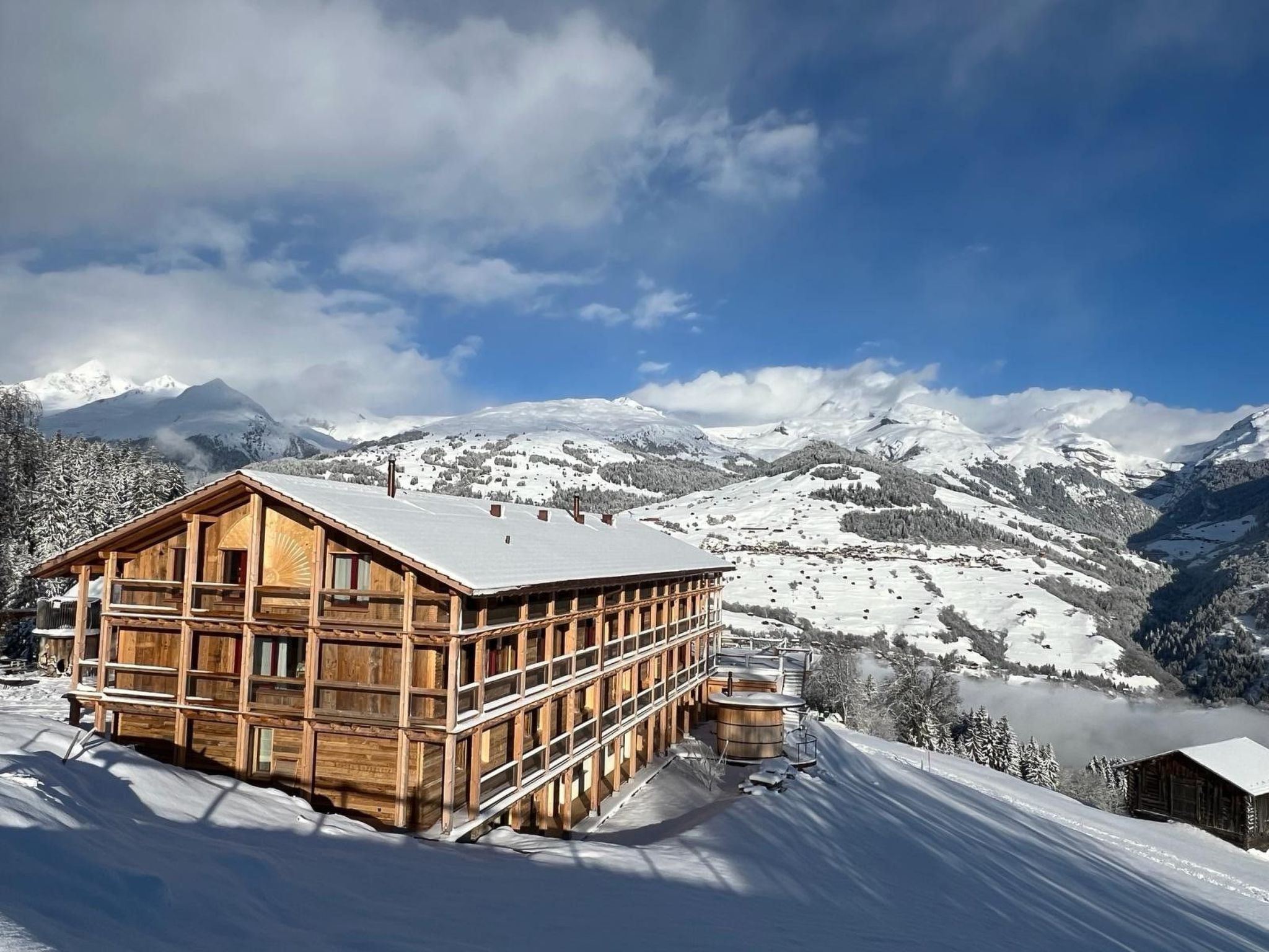 A wooden chalet with a sloped roof, surrounded by snow-covered mountains and a clear sky.