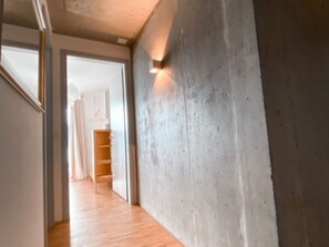 Un pasillo con piso de madera, una pared de concreto con una lámpara, y una puerta que da a una habitación con techo blanco y una lámpara colgante.