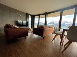 A modern living room with a large window, a flat-screen TV, leather sofas, and a dining area. - Schwalbennest Votteler, Morissen, (Morissen), 43009B by Interhome (Morissen)