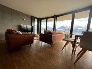 Un salon moderne doté d’un grand téléviseur, de deux canapés, d’une table à manger avec des chaises, et offrant une vue sur les montagnes grâce à de grandes fenêtres.