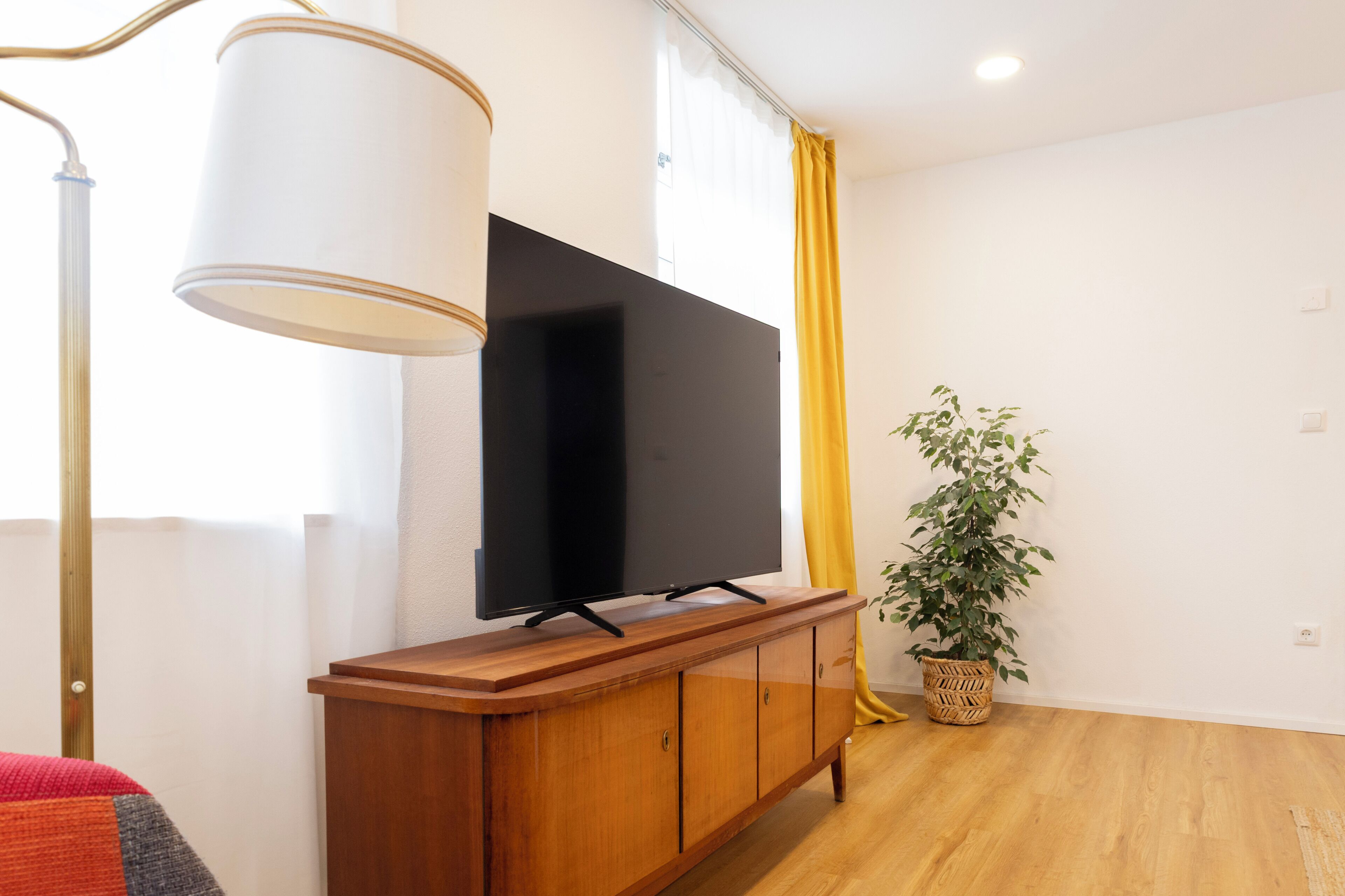 Ein modernes Wohnzimmer mit einem Flachbildfernseher auf einem Holzschrank, einer Stehlampe, einer Topfpflanze und einem gelben Vorhang.