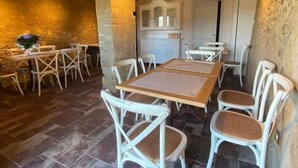 A rustic dining area with a wooden table and white wicker chairs, a stone floor, and a kitchen in the background.