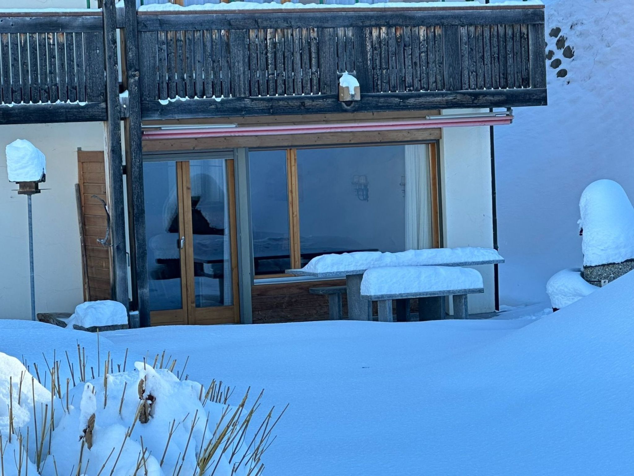 Un bâtiment enneigé doté d’un balcon en bois, d’une porte vitrée et d’un petit espace extérieur aménagé pour s’asseoir.
