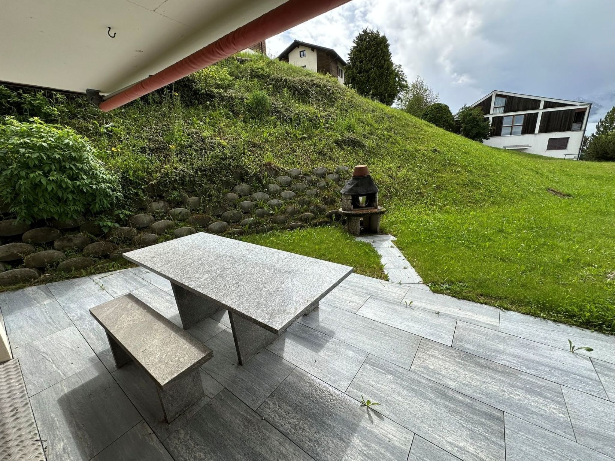 A patio with a stone table and bench, a stone wall, and a grassy hill with a house in the background.