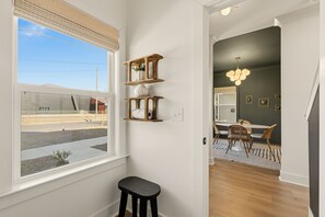 Een kamer met een raam, een kruk, een eethoek met stoelen en een tafel, en een wandplank met decoratieve objecten.
