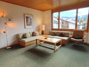 Una sala de estar acogedora con un sofá, una mesa de centro y un sillón. Se aprecia una ventana con vistas a edificios y montañas.