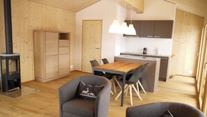 Een modern interieur met een houten vloer, een zithoek met een houten tafel en stoelen, een keuken met donkere kastjes, en een houtkachel.