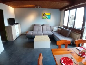 Ein Wohnzimmer mit einer Eckcouch, einem Couchtisch, einem Esstisch mit Stühlen und einem Gemälde an der Wand.