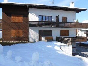 A two-story building with wooden shutters, a balcony, and a snow-covered ground.