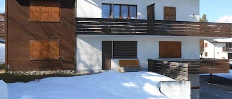 Un edificio de dos pisos con contraventanas de madera, un balcón y el suelo cubierto de nieve.