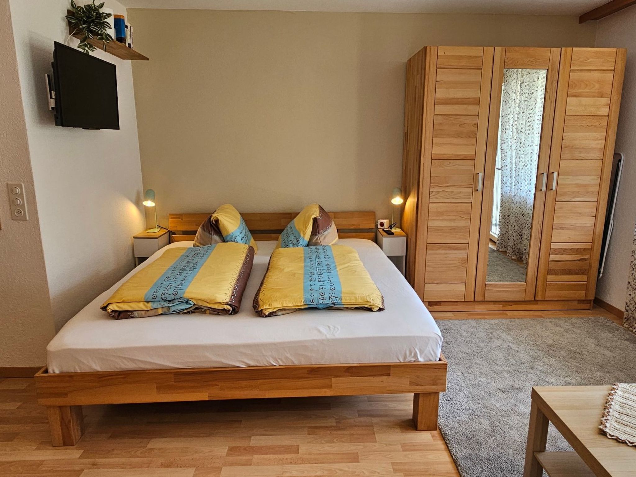 Ein Schlafzimmer mit einem hölzernen Bett, zwei Nachttischen mit Lampen, einem Kleiderschrank und einem an der Wand befestigten Fernseher.