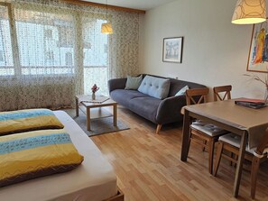 A room with a sofa, a dining table, a small side table, and a bed with patterned bedding.
