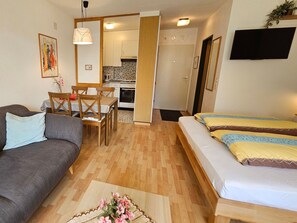 Ein kompakter Wohnbereich mit einer Couch, einem Bett mit gemusterten Betttextilien, einem Esstisch mit Stühlen, einer Küchenzeile und einem an der Wand befestigten Fernseher.