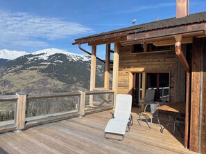 A wooden deck with a view of snow-capped mountains, a covered outdoor seating area with chairs and a table, and a wooden building with a sloped roof.