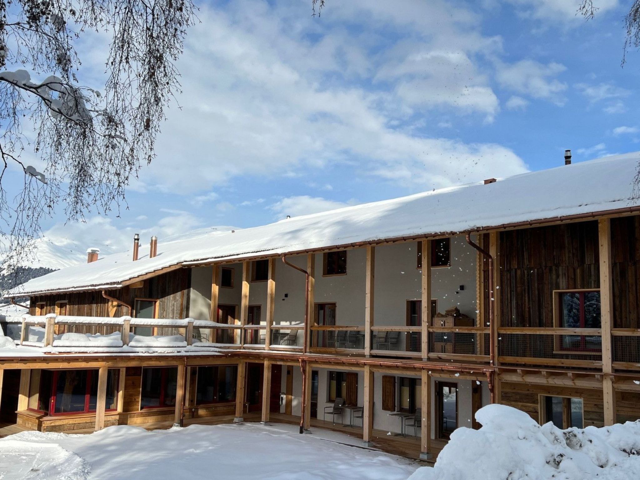 A multi-story wooden chalet with balconies, surrounded by snow-covered ground and mountains.