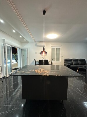 A modern kitchen with a marble countertop, a hanging pendant light, and a black sofa in the background. - Villa Badia (Empuriabrava)