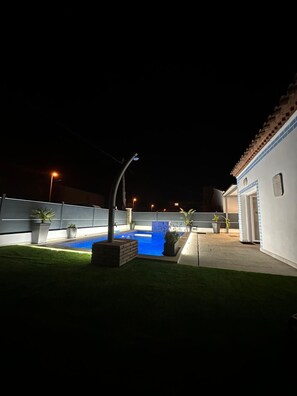 A nighttime view of a swimming pool with surrounding potted plants and a building with a tiled roof. - Villa Badia (Empuriabrava)