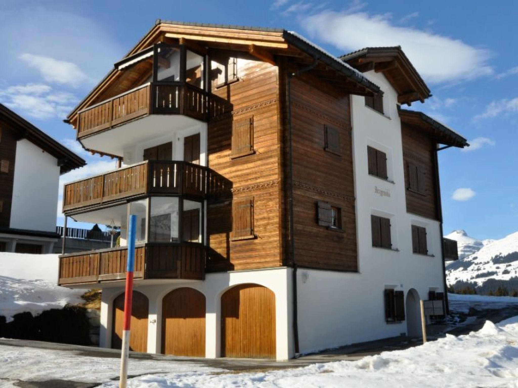 Ein mehrgeschossiges Gebäude mit Holzbalkonen und einem Ski-Hang im Hintergrund, ferner ein klarer Himmel.