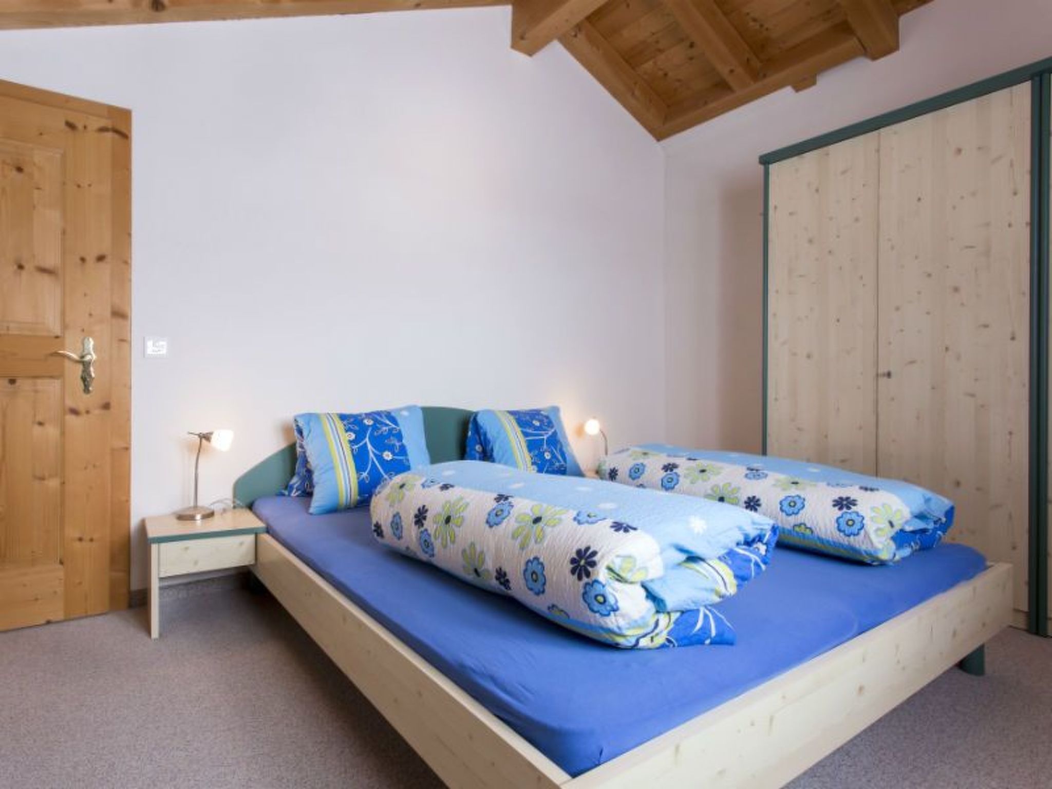 Een enkel bed met een blauwe matras en kussens met bloemenprint, een houten nachtkastje met een lamp, en een houten garderobe met groene randen.