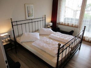 Ein Einzelbett mit Metallgestell, weiße Bettwäsche, ein Nachttisch mit Lampe und ein Fenster mit Spitzenvorhängen.