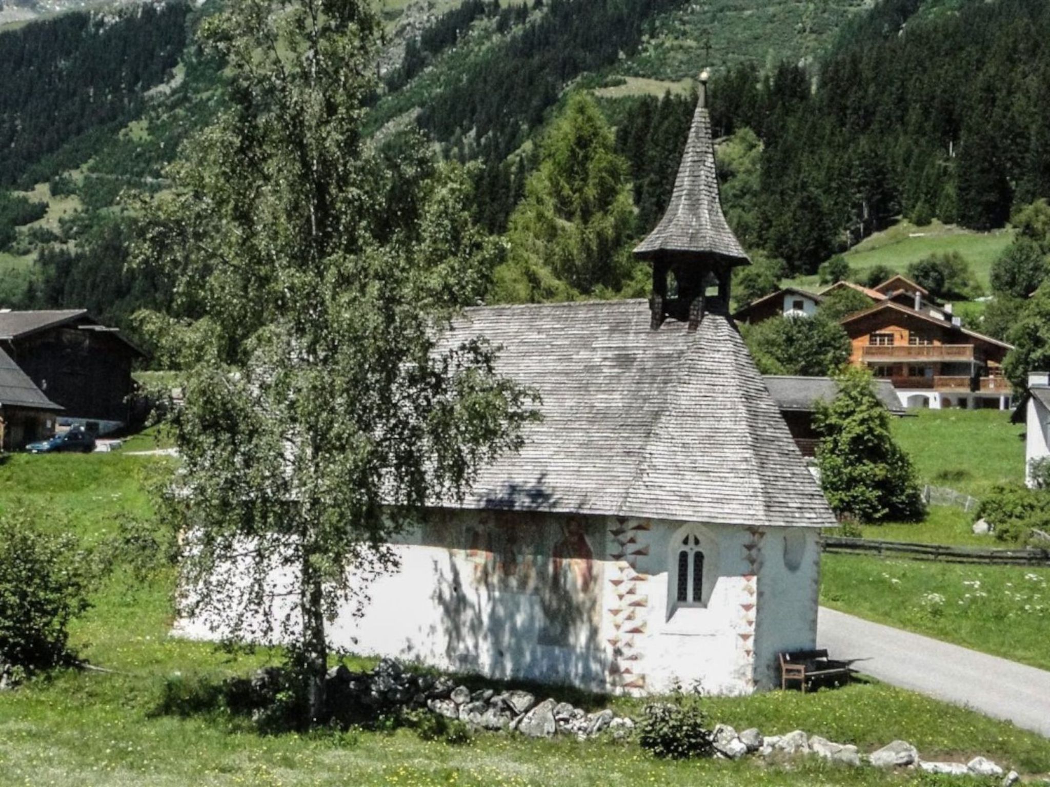 A small white church with a pointed steeple, surrounded by greenery and a few houses in a hilly area.