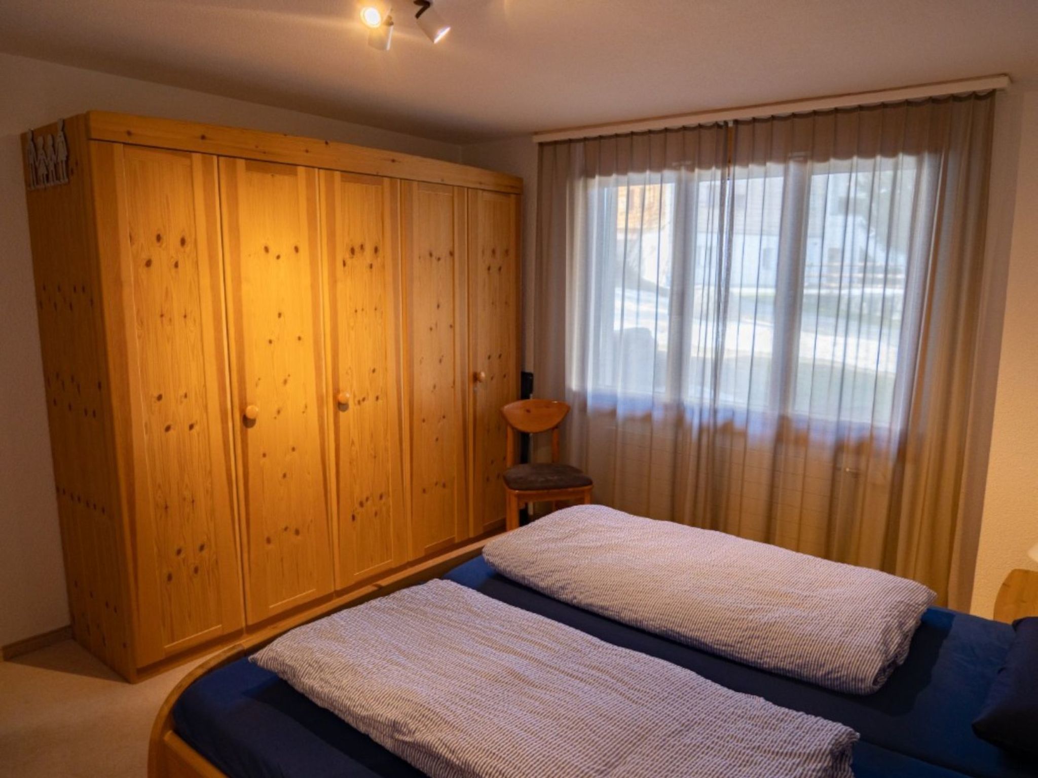Een slaapkamer met een grote houten garderobe, een bed met blauw beddengoed en een raam met dunne gordijnen.