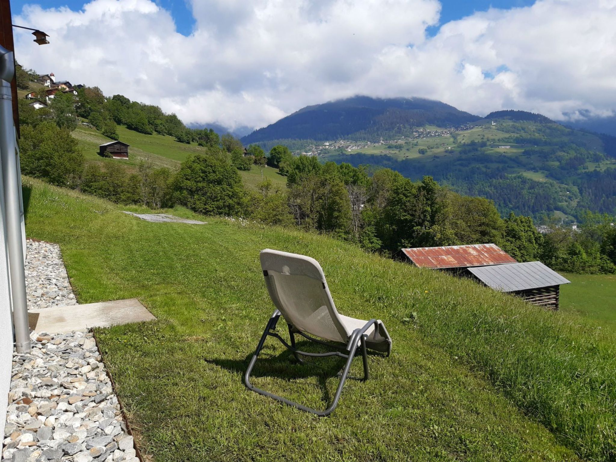 A grassy outdoor area with a metal chair, a small wooden structure, and a view of a mountain range.
