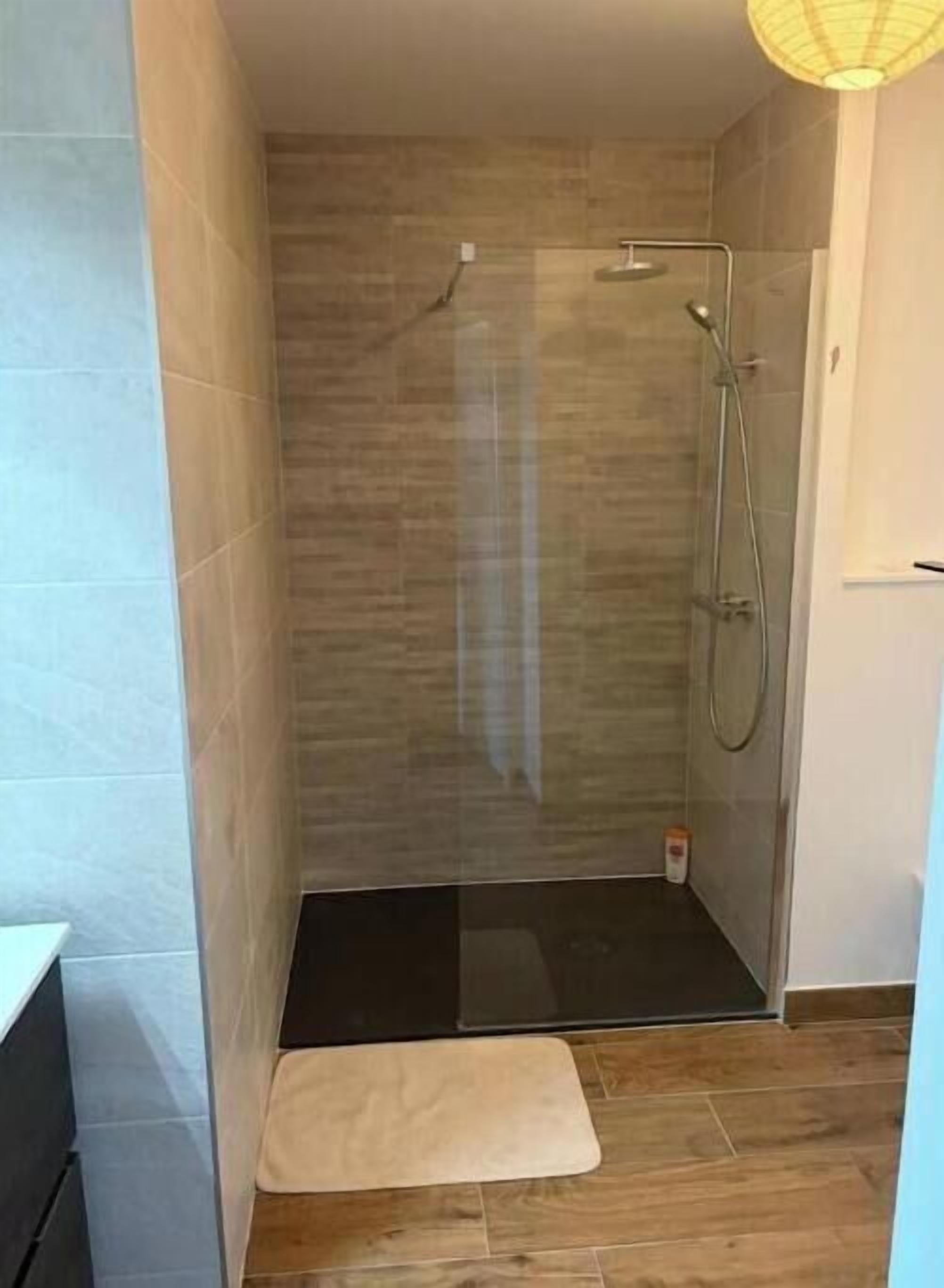 Zona de ducha con puerta de cristal, una pequeña alfombra y paredes de azulejos.