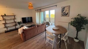 A living room with a brown sofa, a TV, a dining table with chairs, a potted plant, and a ceiling light. - Elegance & Terrace in the heart of the 5 Cantons - Beaches on foot (Anglet)