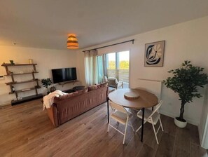 Ein Wohnzimmer mit braunem Sofa, einem Fernseher, einem Esstisch mit Stühlen, einer Topfpflanze und einer Deckenleuchte.