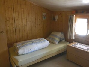 A single bed with a striped pillow and a plain pillow, a wooden dresser, and a window with curtains. - Soldanella Tschuor, Obersaxen-Giraniga, (Obersaxen), 34078B by Interhome (Obersaxen Affeier)