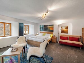 A modern bedroom with a large bed, two white furry chairs, a red sofa, a small table, and a nightstand with a lamp. - Pacifica und Romantica in Cumpadials, (Cumpadials), 76002B by Interhome (Cumpadials)
