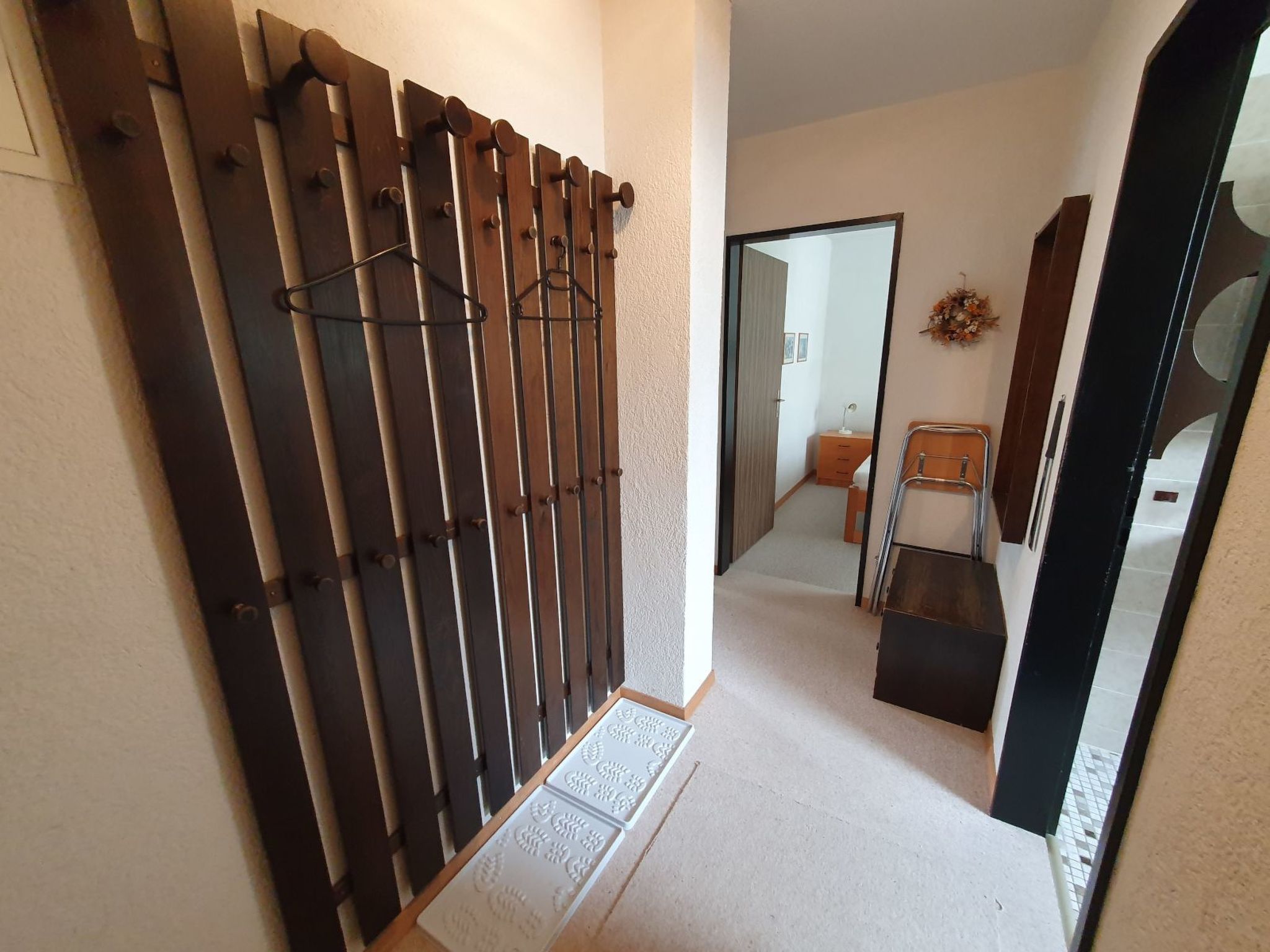 Ein Flur mit einem Holztor, einem kleinen Tisch mit Lampe und einem Spiegel, der ein Zimmer mit Bett und Kommode reflektiert.