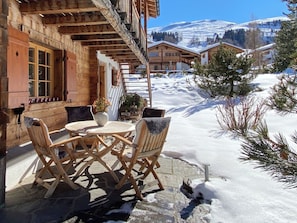 Un chalet in legno con un tavolo e sedie sul patio, una scala in legno e montagne innevate sullo sfondo.