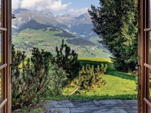 A view through an open doorway with a stone floor, leading to a green landscape with trees and a distant mountain range.