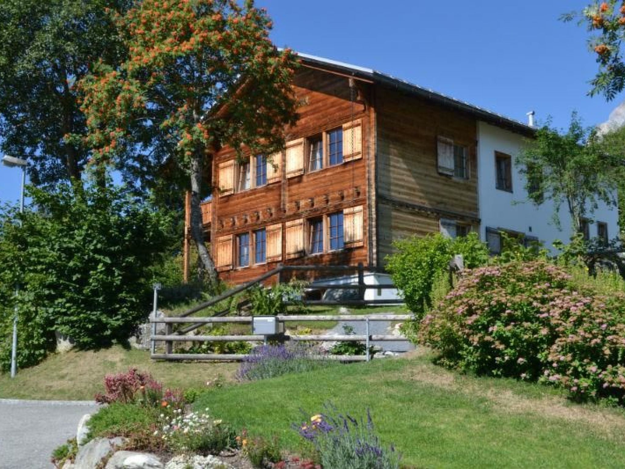 A two-story wooden house with a visible staircase leading to the upper floor, surrounded by lush greenery and flowering plants.
