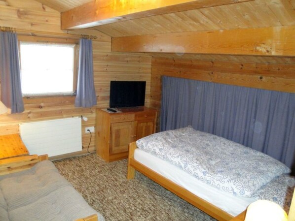 A wooden cabin room with a bed, a television, and a window with curtains. - Gadola, Obersaxen-Meierhof, (Obersaxen), 34069B by Interhome (Obersaxen Affeier)