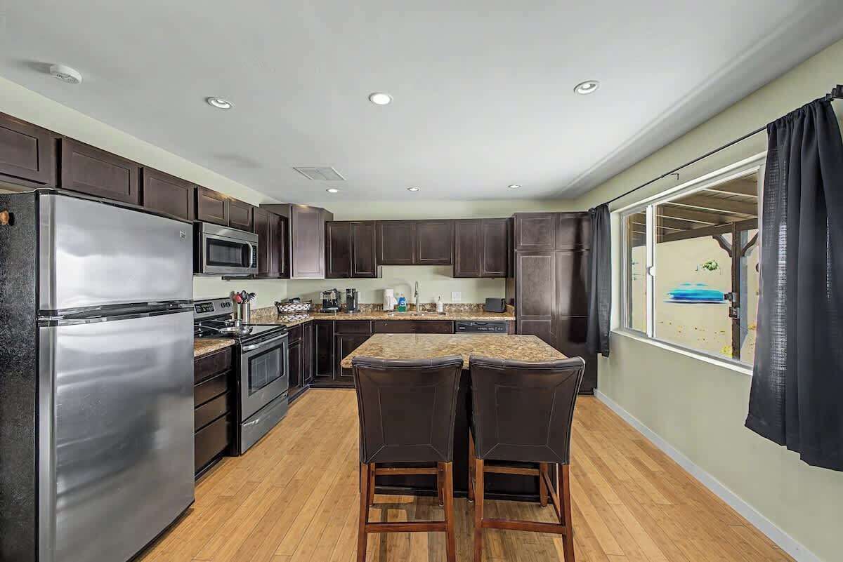 A modern kitchen with dark cabinets, stainless steel appliances, a central island with bar stools, and a large window with curtains.